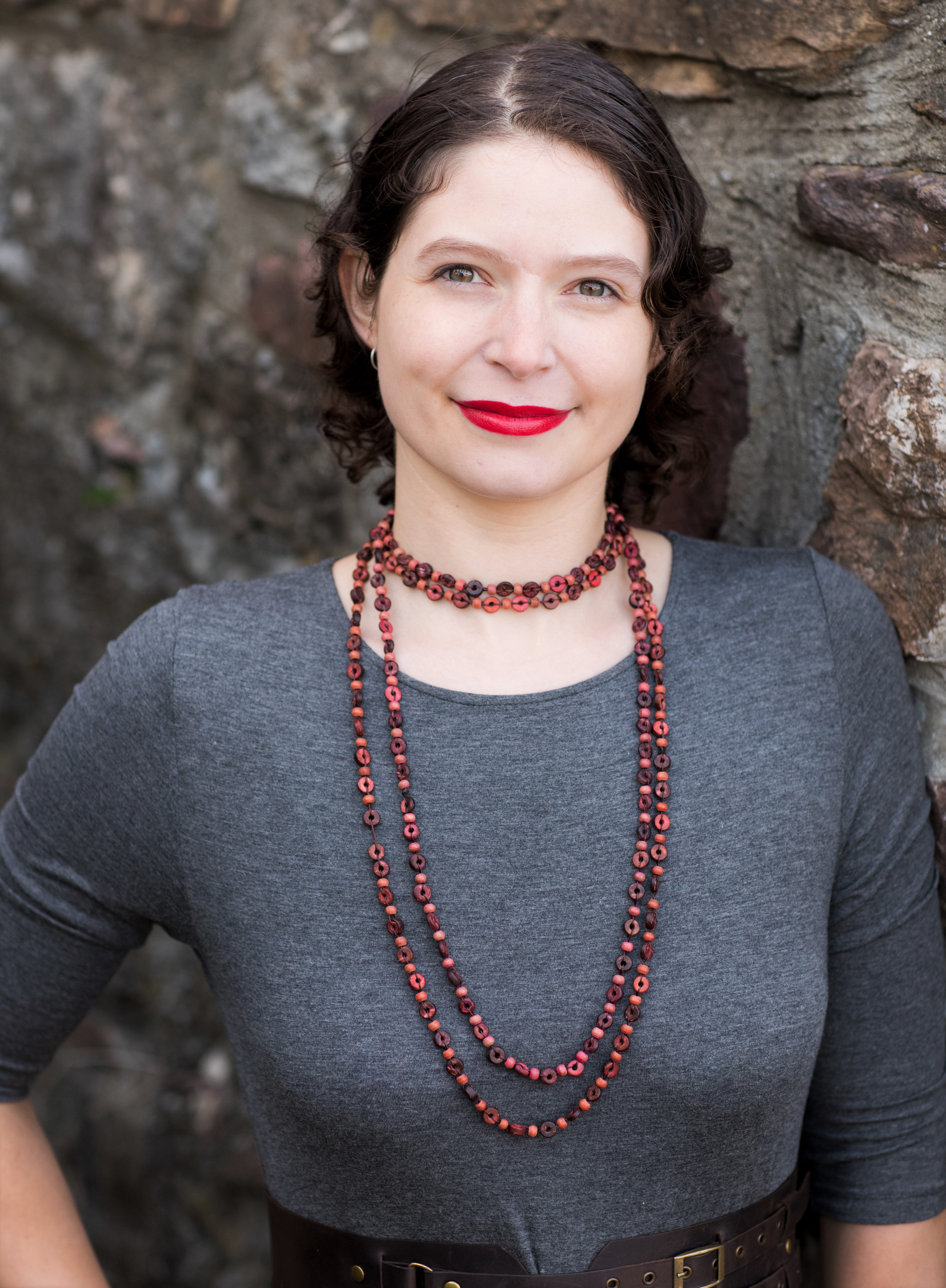 Color photograph of a young woman in a grey dress who is gently smiling at the camera. She is a brunette with curly, shoulder-length hair, pale skin, and hazel eyes. She is wearing red lipstick and a reddish-brown beaded necklace. She is leaning against a masonry wall.